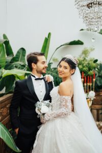 A stylish wedding couple poses in an elegant indoor setting with lush greenery.