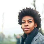 A fashionable black woman with an afro hairstyle in a winter coat poses outdoors in Budapest park.